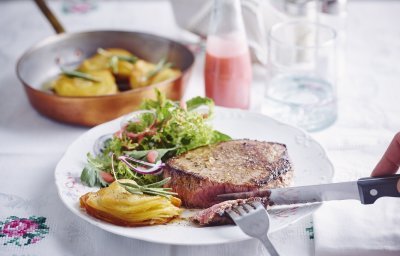 Een hoofdgerecht om U tegen te zeggen. We serveren een malse chateaubriand met millefeuille van aardappelschijfjes en knapperige kruidensalade. De frambozendressing staat langs het bord in een klein glazen flesje. Er staat ook een glas water boven het bord, naast peper en zout.