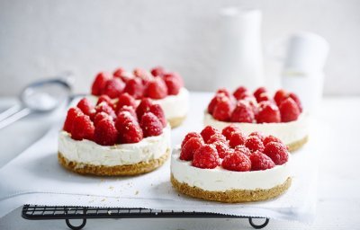 Wij maakten 4 kleine cheesecakes en staan op een wit boterpapiertje. Ze zijn afgewerkt met verse frambozen die een mooi contast geven met het witte van de kaas.