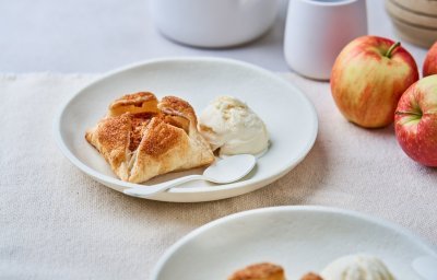 Op een mooi wit bord ligt een appeltaartje van bladerdeeg en een bolletje vanille-ijs. Er liggen ook enkele jonagold appelen langs het gerecht. 