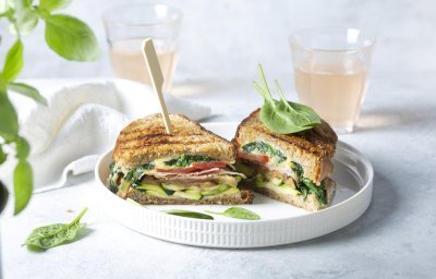 Een aangesneden croque ligt op een wit bord. De croque zit boordevol groentjes, en zit vast met een houten prikker. Achteraan de foto staan 2 glaasje licht roze huisgemaakte limonade.