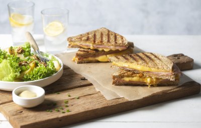 Een krokant gebakken croque monsieur ligt aangesneden op een houten snijplank. Lang staat een salade in een diep wit bord met een potje mayonaise voor te dippen. 