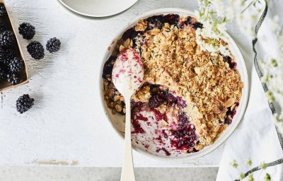 Crumble met havermout en braambessen in een witte ronde ovenschaal. Er is al een portie crumble verdwenen uit de ovenschaal en er liggen enkele braambessen langs. Om nog wat extra sfeer toe te voegen zijn er witte fijne bloemetjes rechts in beeld.