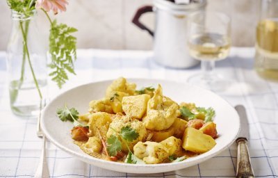 Heerlijke goudkleurige curry geserveerd in een diep wit bord met bloemkool en aardappelen. Er ligt bestek langsheen het bord en glaasje witte wijn. Ook zien we een vaasje met roze bloem en varentakje. 