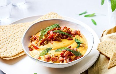 Er staat een diep bord op tafel, waarin de gemixte butternut met geitenkaas ligt. De dip is afgewerkt met stukjes gebakken spek, salie, noten en kruiden. Hij is prachtig oranje. Wij serveren hem met lange crackers. 
