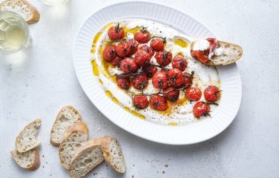 Een groot ovaal bord is gevuld met de romige yoghurt waarop de dieprode kerstomaatjes bovenop liggen. Er liggen enkele sneetjes stokbrood naast om te dippen. Er staan ook twee glaasjes witte wijn in beeld. 