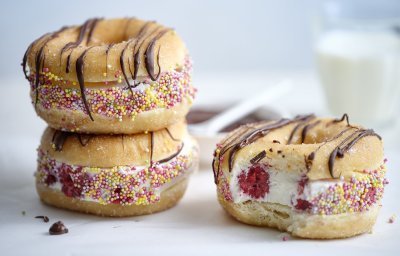 Drie donuts liggen op een witte stenen snijplank. De donuts zijn doormidden gesneden en gevuld met yoghurtijs met rode framboosjes, gerold door kleurrijke sprinkles en afgewerkt met gesmolten chocolade. In de achtergrond staat nog een glaasje melk.