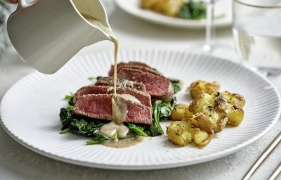 Een wit bord staat op tafel met hierop de heerlijke krokante aardappel en de saignant gebakken filet pur op een bedje van spinazie. Er komt een sauskannetje in beeld die saus over het vlees giet. De tafel is gedekt voor twee met de perfecte maaltijd.