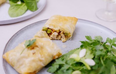 Een heerlijk cadeautje van filodeeg gevuld met stukjes kip, zoete aardappel, erwtjes en grilkaas ligt op een licht blauw bordje. We serveren het met een groen slaatje en lente-uitjes. Het filodeegpakketje is doormidden gesneden. 