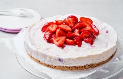 Een heerlijke cheesecake staat op een glazen serveerschaal. Dit is niet zomaar een cheesecake, maar eentje die diepgevroren is. Perfect voor een koel dessertje op een zomerse dag. Er liggen enkele verse aardbeien op de taart om het fruit extra in de verf te zetten, want aardbeien zijn gewoonweg de toppers van het zomerfruit. 