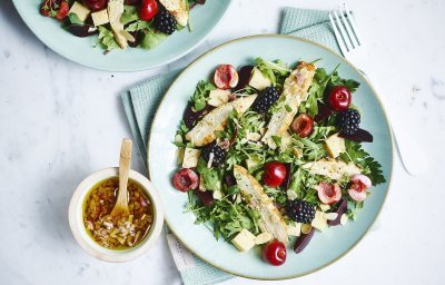 Een gemengde salade met sla, kersen, bramen en gekookte kipfilet gedresseerd op een lichtblauw bord. De dressing is er naast gezet in een keramieke potje met houten lepeltje in, de dressing bestaat uit sjalot, rode wijazijn, olijfolie en peper en zout. 