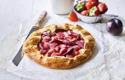 Een klaargemaakte galette staat op een houten witte tafel. Hij is prachtig goudgeel gebakken en het fruit oogt zoet-rode tinten. Er liggen nog wat aardbeien langs, alsook een paarse pruim.