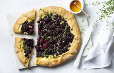 Galette met bramen, blauwe bessen en Belgische brie ligt aanhesneden op het aanrecht. Vergezeld met een potje gouden honing en een tijmplantje. 