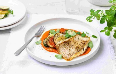 Gebakken rog met ratatouille en paprikasaus geserveerd op een wit groot bord. Het vork ligt op het bord en verse kruiden staan langs deze hoofdmaaltijd. 