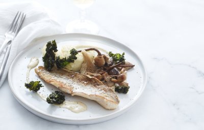Een gebakken visje op vel ligt op een wit bordje. Hij is afgewerkt met gebakken boschampignons en krokante stukjes boerenkool. Wij serveren hem met knolselderpuree en een glaasje witte wijn.