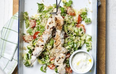 Grote witte bakplaat staat op tafel met eerst een quinoaslaatje en groentjes gepresenteerd.  Hierop liggen de gegrilde brochettes van kip met een dressing van yoghurt. 