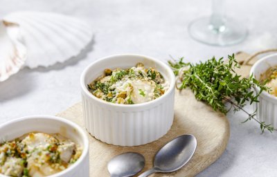 Gegrilde sint-jakobsvruchten gepresenteerd in kleine witte ovenschaaltjes met een crumble van crackers, kaas en verse kruiden. Ze staan op een houten plankje met kleine lepeltjes en verse tijm naast. 