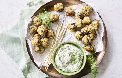 Op het aanrecht staat een houten ronde serveerschaal met een wit boterpapiertje op. Hierop liggen kleine gehaktballetjes met groene stukjes in van de courgette en spinazie. De dip op basis van tzatziki staat ook op de schaal. Het gerecht is afgewerkt met enkele takjes dille. 