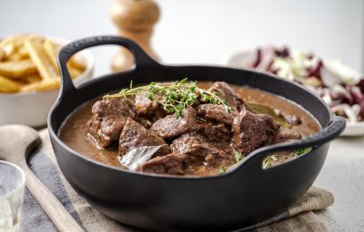 Er staat een zwarte sudderpot op tafel met Gentse stoverij, in de achtergrond zijn er frietjes te zien en een witloofslaatje.