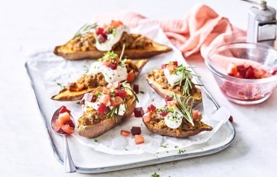 Op het aanrecht staat een rechthoekige porseleinen serveerschaal met hierop de gevulde zoete aardappelen. Het gerechtje heeft een kleur waar je blij van wordt! Het witte toefje van de zure room, blokjes rode biet en tomaat en afwerken doen we met een frisgroen takje rozemarijn. Een prachtig gerecht in smaak en kleur!