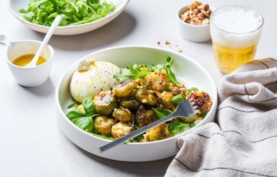 In een diep wit bord liggen de geroosterde spruitjes op een bedje van rucola en basilicum. Hierlangs ligt de burrata. Er is al wat vinaigrette gesprenkeld op de groenten en burrata.