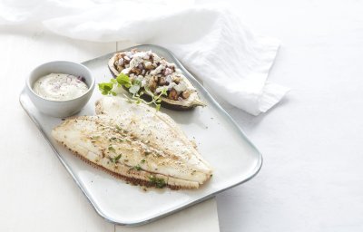 Een gebakken schartong ligt op een rechthoekig wit bord. De halve aubergine ligt naast de gebakken vis, hij is gevuld met stukjes knolselder, peer en walnoten. Het gerechtje is afgewerkt met verse gehakte kruiden en een lepeltje zure room.