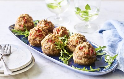 Op een blauw schaaltje liggen gevulde champignons. Ze zijn geserveerd met enkele blaadjes rucola en een stappeltje bordjes met mini-vorkjes om het hapje proper te eten. 