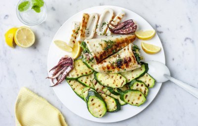 Groot wit bord met gegrilde courgetteschijfjes en gevulde inktvis. Er zijn partjes citroen toegevoegd die zorgen voor het lentegevoel in het beeld samen met de pastelgele servette. 