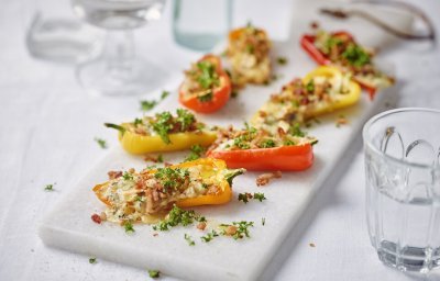 Gele en oranje snackpaprika's liggen op een witte stenen snijplank. De paprika's zijn gevuld met kaas, noten en crumble van spek. Ze zijn afgewerkt met verse peterselie. 