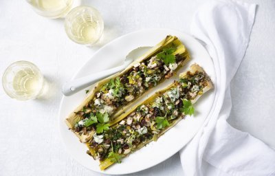 Twee gevulde preien liggen op een groot bord. Er staat een glaasje witte wijn bij en een wit servet.  De prei is gevuld met champignons, noten en blauwe kaas.