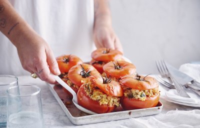 Zes gevulde rode tomaten in een ovenschaal zijn aan tafel gebracht door een vrouw in witte t-shirt. Ze schept alvast eentje op voor zichzelf.