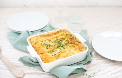 Een gratin staat op tafel onder een lichtblauwe handdoek. Hij is klaargemaakt in een vierkante witte ovenschotel en afgewerkt met verse tijmblaadjes.