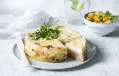 Een hartige cheesecake smaakt heerlijk als lunch of avondmaal. Door het tomatenslaatje krijg je de frisse toets die de cheesesmaak verfrissen. De cheescake ligt aangesneden op een groot rond bord, het slaatje staat langs het bord en een karaf water met takje munt zie je achteraan in de foto. 