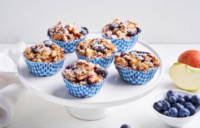 Op een taartenserveerschaal staan 6 muffins in blauw-witte muffinpapiertjes. Er ligt nog een appel langs en blauwe bessen in een potje. 
