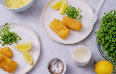 Twee witte bordjes staan op een licht grijze tafel, op elk bordje liggen twee kaaskroketten met partjes citroen en verse tuinkers. 