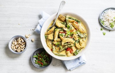 Er staat een grote witte serveerschaal op tafel gevuld met de goudgele kip curry. Je ziet de groene peultjes en rode chili duidelijk. Langs het gerecht staan nog extra noten, gesneden lente-uitjes en een bordje rijst. 