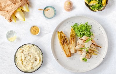 Gestoofd witloof ligt op een wit bord, samen met de kipfilet en mosterdsaus. De groene rucola zorgt voor een frisse kleur in dit gerecht. 