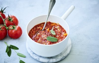 Een klassieke tomaten die is klaargemaakt in een witte kookpot. De tomatensaus zit boordevol verse groenten en tomaten, natuurlijk. Er liggen enkele blaadjes salie op de saus en een verse tros tomaten naast de pot op het marmere aanrecht.
