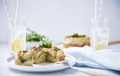 Kleine quiche van brood met broccoli en paprika op wit bordje met houte bestek langs het bord. Er staan ook twee waterglazen met een schijfje citroen en kartonnen rietje in.
