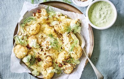 Puree van knolselder is uitgeschept op een groot lichtbruin bord. Hierop zijn de geroosterde bloemkoolroosjes geserveerd met wat drizzle van de groene kruidendressing, noten en verse dille. De overige kruidendressing is in een witte potje en staat langs het bruine bord, samen met nog wat gehakte noten in een ander potje. Alles staat op een een plaid in een blauwgrijze kleur.