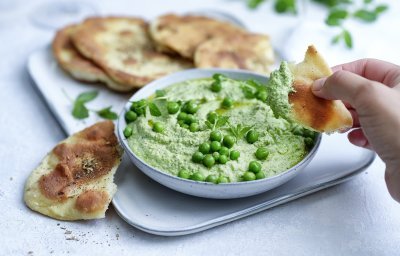 Krokant gebakken platbrood ligt op een serveerschaaltje waarbij een dip van erwten en roomkaas in geserveerd. De dip is gemixt en heeft een mooie groene kleur, om af te werken zijn er nog enkele verse erwtjes bovenop gestrooid. Een hand neem alvast een stukje platbrood om te dippen in gezonde groentedip. 