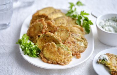 Krokante courgetteplakjes liggen op een bordje, afgewerkt met enkele blaadjes peterselie. De aioli staat langs het bordje.
