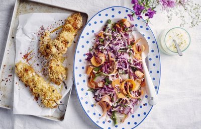 Krokante kipbrochette ligt nog de sidderen op een ovenplaatje. Daarnaast staat een kleurrijk groot bord met een salade op basis van rode kool, wortel en komkommer. De dressing op basis van yoghurt staat langs de salade.