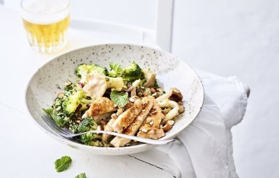 Een rijkgevulde quinoasalade boordevol verse groenten en kruiden zoals broccoli, paddenstoelen, rode ui, peterselie, koriander en munt. Bovenop de quinoa werd het varkensvlees geplaatst in schijfjes. Er staat een witbier bovenaan het bord, op de tafel.