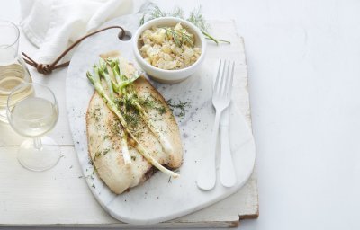 De gebakken vis ligt op een stenen witte snijplank, waar de gigrilde lente-ui bovenop ligt. De Macédoine van venkel en krieltjes is opgediend in een apart wit potje. Er ligt wit bestek langs, met een glaasje witte wijn en een linnen servette. Het gerechtje is afgewerkt met verse dille.