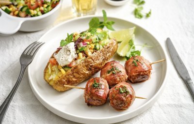 Een gepofte aardappel ligt op een rond wit bord. Hij is gevuld met maïs, tomatenstukjes, komkommer en een toefje zure room. Hierlangs liggen 2 spiesjes met gelakte balletjes op geprikt. 