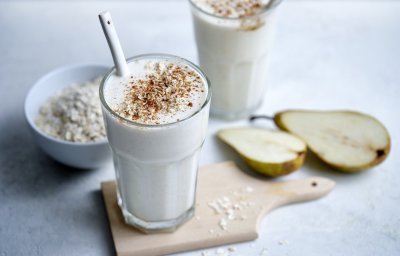 Twee klaargemaakte milkshakes staan op het lichtgrijze aanrecht te pronken. Er ligt een peer langs die doormidden gesneden is en wat havermoutvlokken gemorst. De shakes zijn afgewerkt met wat kaneelpoeder. 