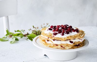 Grote ronde millefeuille met kersen en verse roomkaas. Deze staat op een witte taartschaal op een bleke houten tafel. Op tafel ligt ook een takje groen voor extra sfeer. 