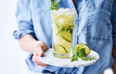 Een heerlijk frisse mocktail van Jupiler 0.0 en peach ice tea. Geserveerd met frisse limoenschijfjes in het glas. De vrouw draagt een blauw jeanshemd en staat mee in beeld.