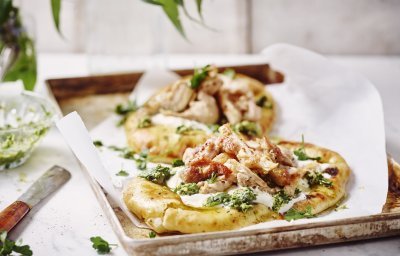 Twee naanbroodjes liggen op een metalen ovenplaat. Ze zijn belegd met yoghurt, stukjes gebakken kip en afgewerkt met salsa verde. De ovenplaat staat op een witte houten tafel.