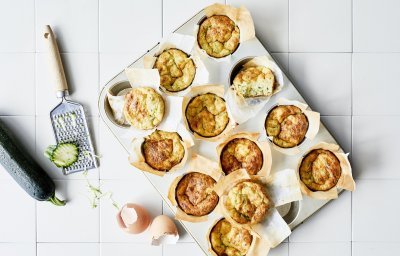 Ontbijtmuffins met courgette, yoghurt en Belgische kaas in een grote muffinvorm. Klaar om eruti te halen en op te eten. Naast de bakvorm ligt een verse courgette en lege eierschalen voor de sfeer.